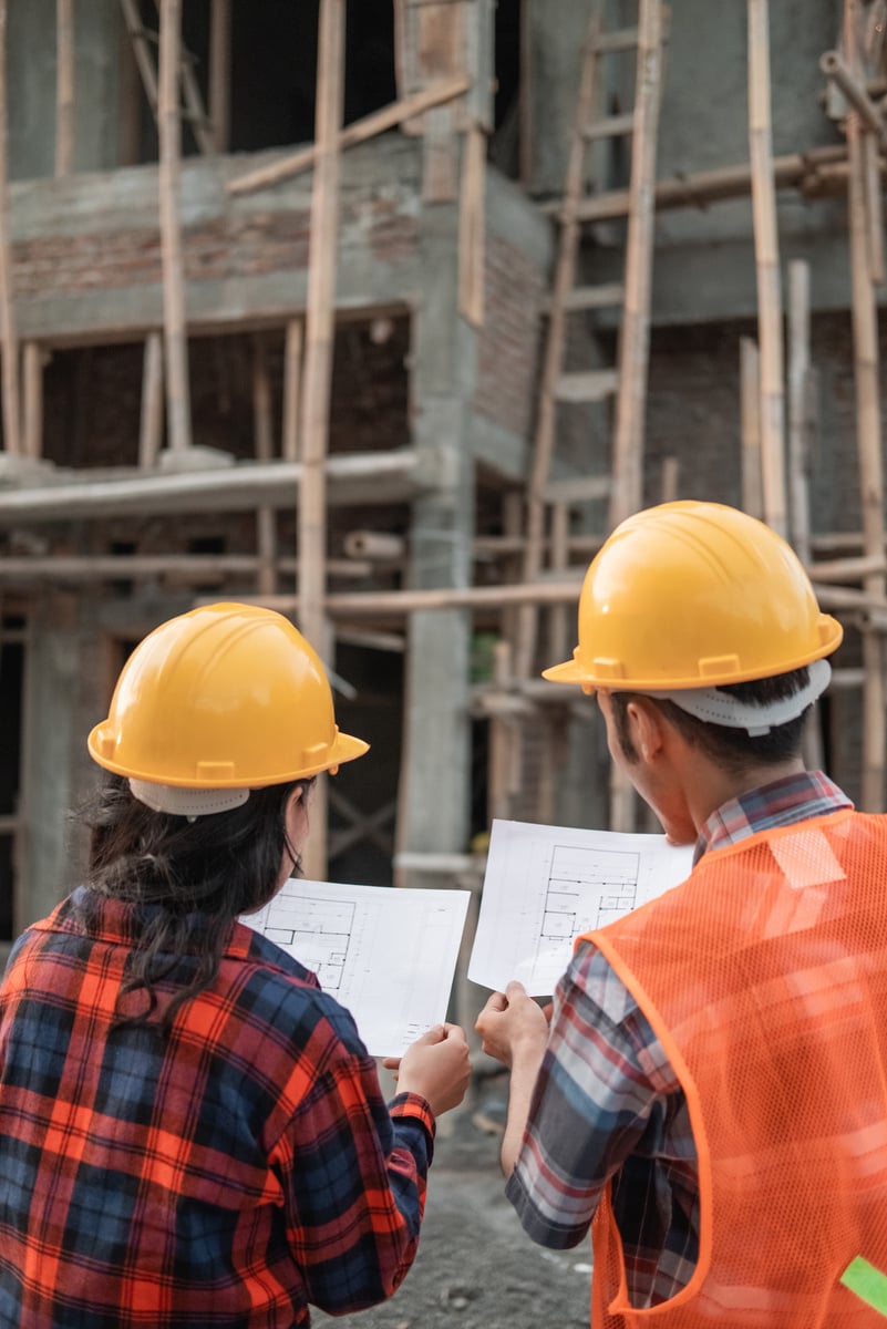 View from behind of Male and Female Asian Contractors Standing Wearing Safety Helmets Holding the Site Plan Building